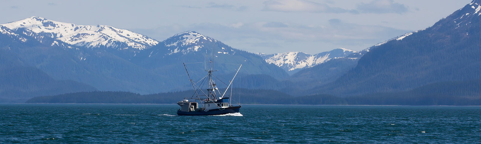 Small fishing boat on deep blue water with a backdrop of snow-capped mountains and a cloudy sky